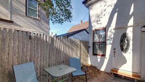 A patio with two chairs and a table, a wooden fence, and a house with a white door and window. - Cozy Studio in the Heart of Bar Harbor (Bar Harbor)