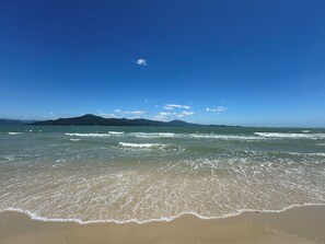 A beach with clear water, gentle waves, and distant mountains under a blue sky. - Apartamento Canajurê com Piscina Floripa (Florianópolis)