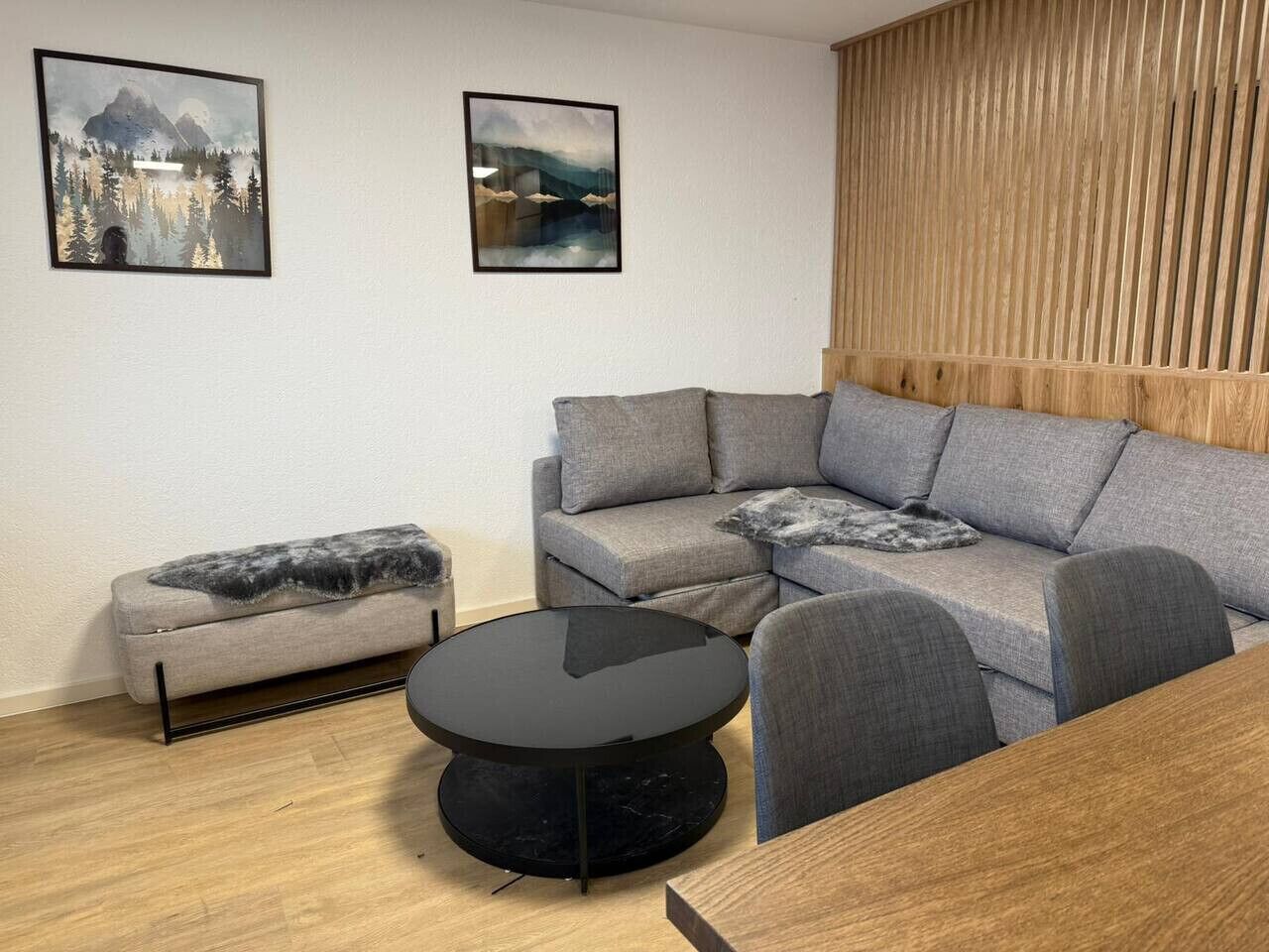 Un salon moderne avec un canapé d’angle gris, un petit canapé gris, une table basse ronde noire et un mur d’accent en bois.