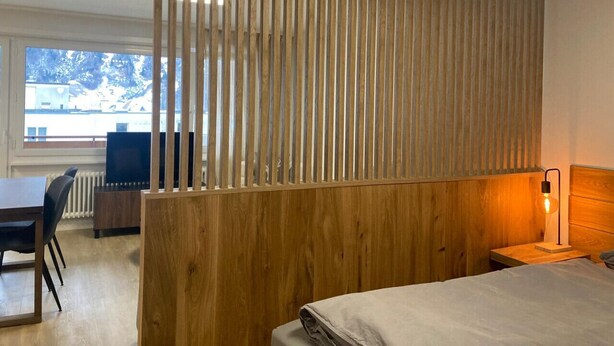 Habitación de hotel moderna con separador de madera, cama, mesita de noche con lámpara, escritorio con silla y vistas al exterior.