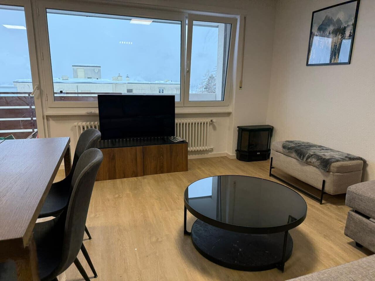 A modern living room with a wooden dining table, a black sofa, a round glass coffee table, and a flat-screen TV on a wooden stand.