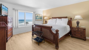 A bedroom with a large bed, a dresser, a nightstand, a window with a view of the ocean, and a painting on the wall. - Sandpiper 611 (New Smyrna Beach)
