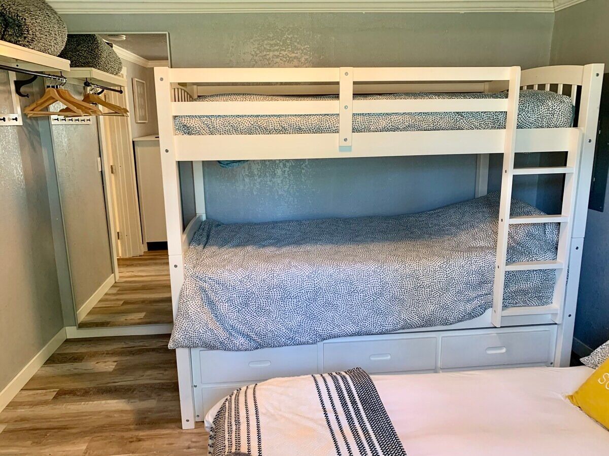 Een stapelbed met een bedspread met patroon, een klein lade en een kledingrek met kleding.