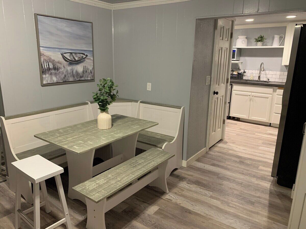 Un comedor con una mesa y bancos, una planta sobre la mesa y una cocina al fondo.