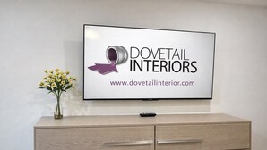 A flat-screen TV mounted on a wall displaying 'Dovetail Interiors' logo and website. Below the TV, there's a wooden cabinet with drawers. - Downtown Clermont Boutique Garden Guest House (Clermont)