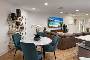 Uma sala de estar moderna com uma mesa redonda, cadeiras verde-água, um sofá marrom e uma grande pintura com tema praiano na parede.