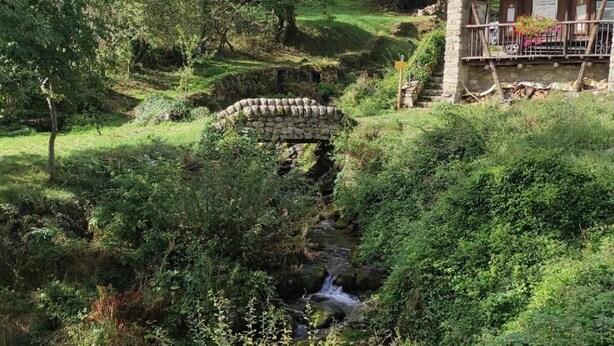 A rustic stone building with a wooden deck, a stone bridge, and a stream in a lush green valley.