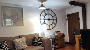 A living room with a large wall clock, a sofa with patterned cushions, a wooden cabinet with books, and a dining area with a chair. - The Old Hanging Gate Inn built in the early 1800s as a former coach house inn. (Telford)
