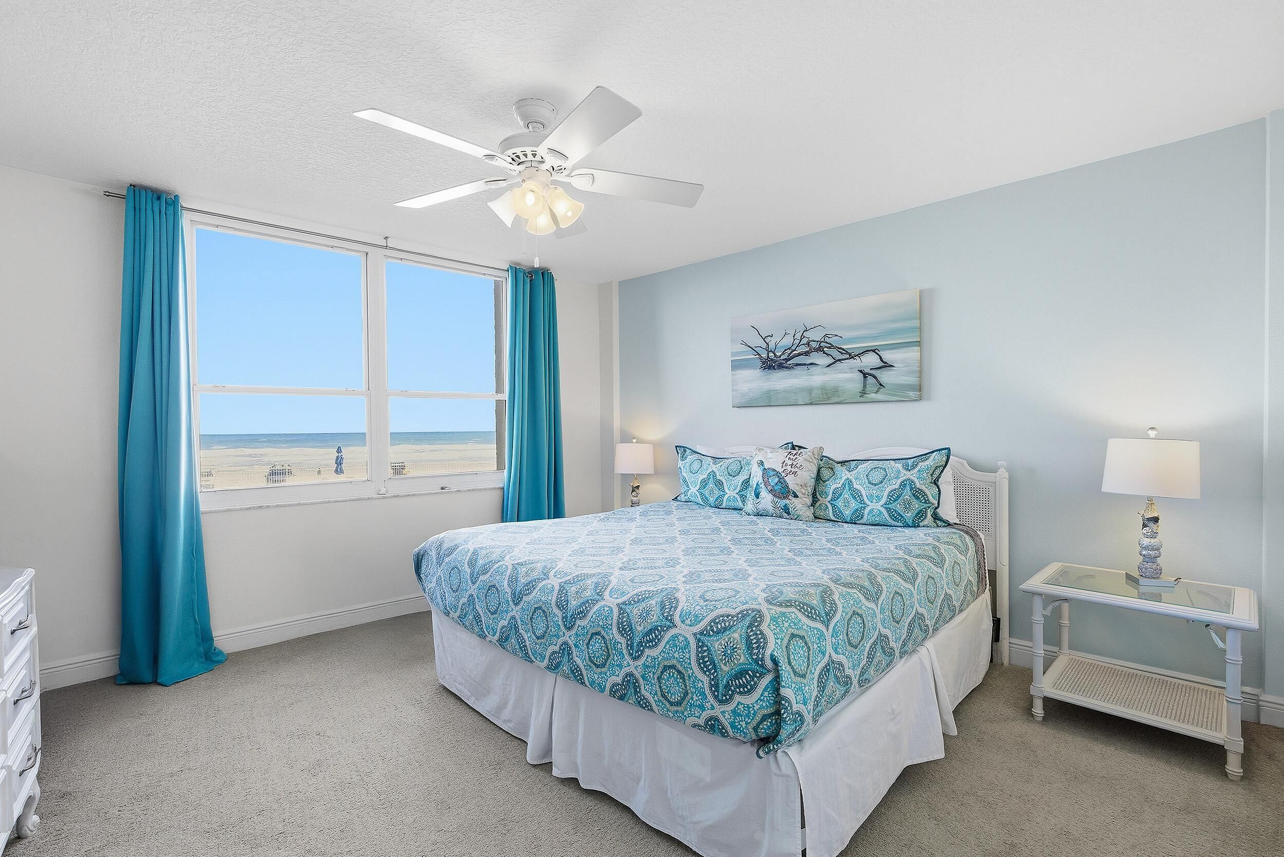 A bedroom with a bed, a ceiling fan, a window with curtains, a bedside table, and a wall-mounted lamp.