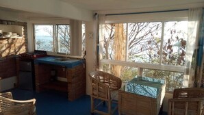 A cozy room with a brick fireplace, a dining table with chairs, and large windows offering a view of trees and a body of water. - Casa Wow "Blue Apartment," where you will wake up to incredible views! (Puerto Ángel)