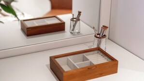 Une boîte en bois avec des compartiments, posée sur une surface blanche, un miroir qui reflète la boîte, et un petit récipient en verre contenant des pinceaux.