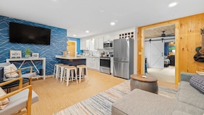 Una sala de estar moderna con sofá, una mesa de centro y televisor. Hay una cocina con refrigerador, microondas y estufa.