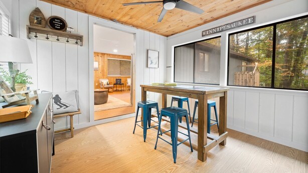 Una habitación acogedora con techo de madera, una mesa de comedor con taburetes azules, una ventana con vistas a árboles y un letrero que dice "Winnipesaukee".