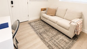 Ein Zimmer mit einem beigen Sofa, einem gemusterten Teppich, einer weißen Tür und einem Fenster mit Jalousien.