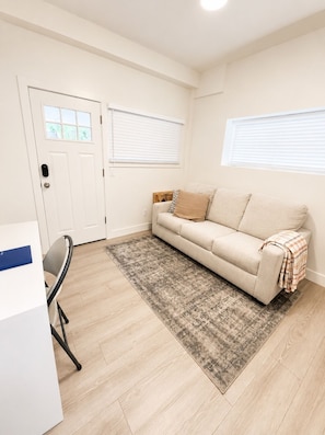 A room with a beige sofa, a patterned rug, a white door, and a window with blinds.