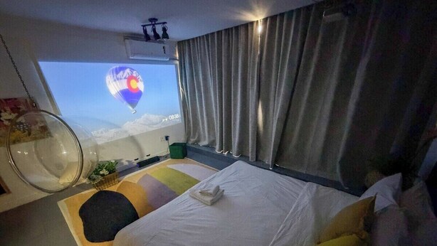 Ein Hotelzimmer mit einem Bett, einem Fenster mit Vorhängen, einem an der Wand montierten Projektor und einem kleinen Tisch mit einer Pflanze.