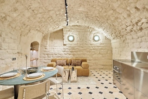 A cozy room with a stone wall, a round dining table set with plates and glasses, a sofa with cushions, and a kitchen area with a stainless steel appliance. - Sublim new Triplex on Place des Vosges (Paris)