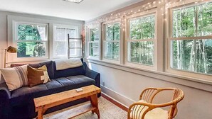A cozy living room with a dark-colored sofa, a wooden coffee table, and a wicker chair. Large windows with white frames allow natural light and views of greenery outside. - Vermont Escape in Ludlow Village - Okemo (Ludlow)