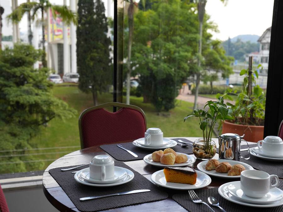 Mesa lista con platos, vasos y un postre, con vistas a un paisaje verde con árboles y un edificio al fondo.