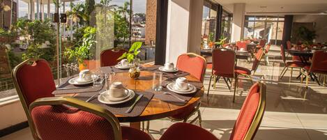 A dining area with a view of a cityscape, featuring a table set with plates, cups, and a vase with a plant.