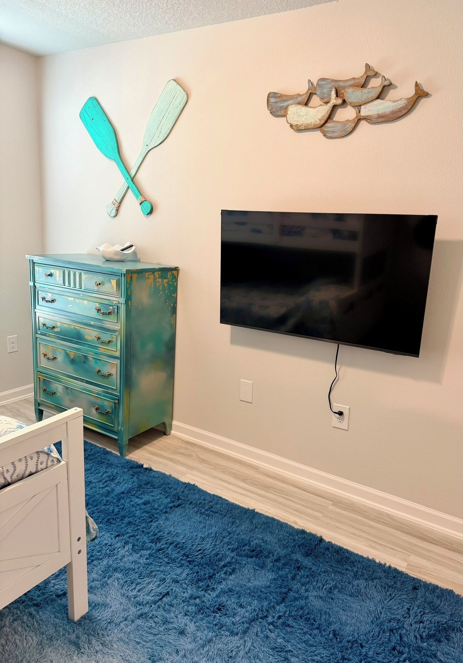 A room with a television mounted on the wall, a blue shaggy carpet, a white dresser, and wall decorations including two paddles and a fish.