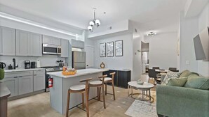 Une cuisine moderne avec des meubles gris, un îlot central doté de tabourets, un coin repas avec un canapé vert et un salon agrémenté d’un tapis à motifs.