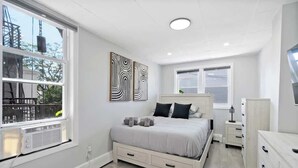 Ein modernes Schlafzimmer mit einem großen Bett, Nachttischen und einer Kommode. Zwei Fenster mit Jalousien sind vorhanden, und eine Heizung ist an der Wand zu sehen.