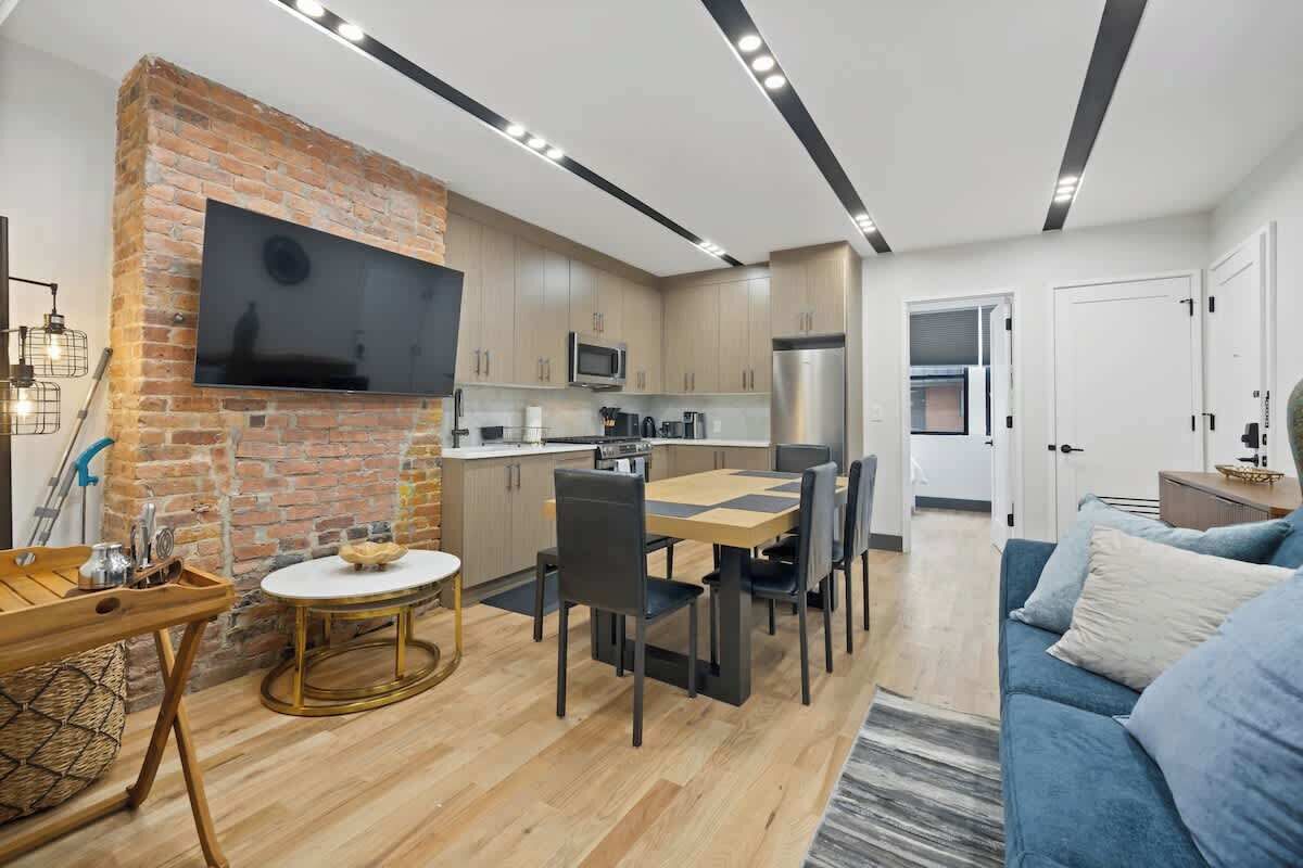 Une cuisine moderne avec un mur d’accent en briques, un coin repas avec une table ronde et des chaises, et un salon avec un canapé bleu.