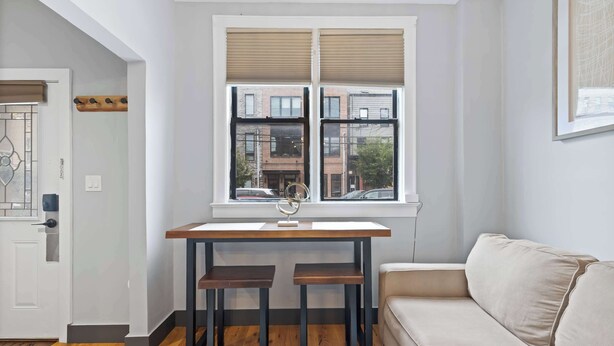 A modern living room with a dining table, two chairs, a sofa, and a window with blinds. - Modern Studio < 20 Min to NYC w/ Fast WiFi (Jersey City)