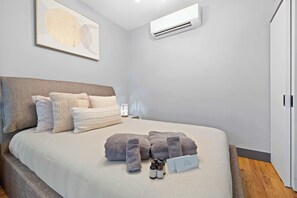 A neatly made bed with a grey blanket and pillows, a bedside table with a lamp, and an air conditioning unit mounted on the wall. - Modern Studio < 20 Min to NYC w/ Fast WiFi (Jersey City)