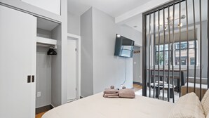 A modern bedroom with a large bed, a TV mounted on the wall, and a balcony with a view of the city. - Modern Studio < 20 Min to NYC w/ Fast WiFi (Jersey City)