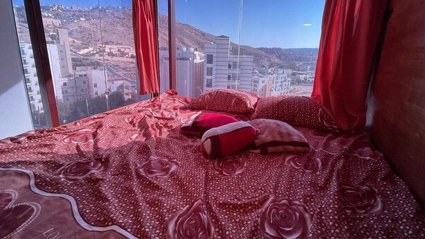Un dormitorio con una cama grande, ropa de cama roja y vistas de un paisaje urbano a través de amplios ventanales.