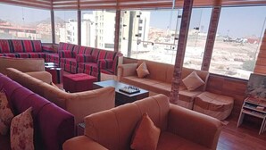 Amplia sala de estar con varias zonas de estar, incluyendo sofás y sillones, dispuestas alrededor de una mesa de centro. Grandes ventanales ofrecen vistas de los edificios y el paisaje circundante.