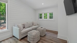 A room with a light-colored sofa, two woven ottomans, a window with a view of trees, and a mounted flat-screen TV.