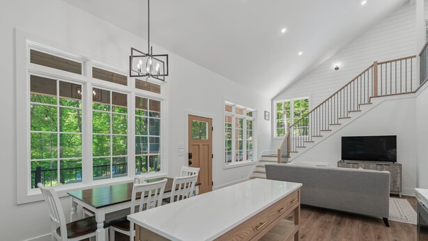 A modern kitchen with a dining area, large windows, a staircase, and a TV.