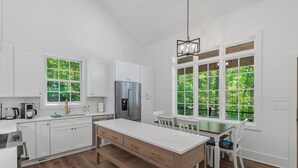 A modern kitchen with white cabinets, a stainless steel refrigerator, a wooden dining table, and large windows with a view of greenery.