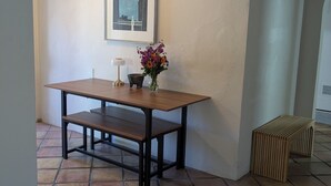Un comedor con mesa de madera, bancos, un jarrón con flores, un cuadro en la pared y suelo de baldosas.