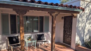 Un porche cubierto con dos sillas y una mesita, una puerta con detalles decorativos y una ventana con contraventanas.