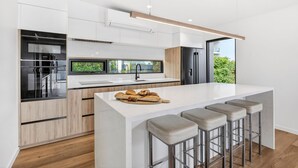 A modern kitchen with a central island, bar stools, and a view of greenery outside. - Teremoana (Lorne)