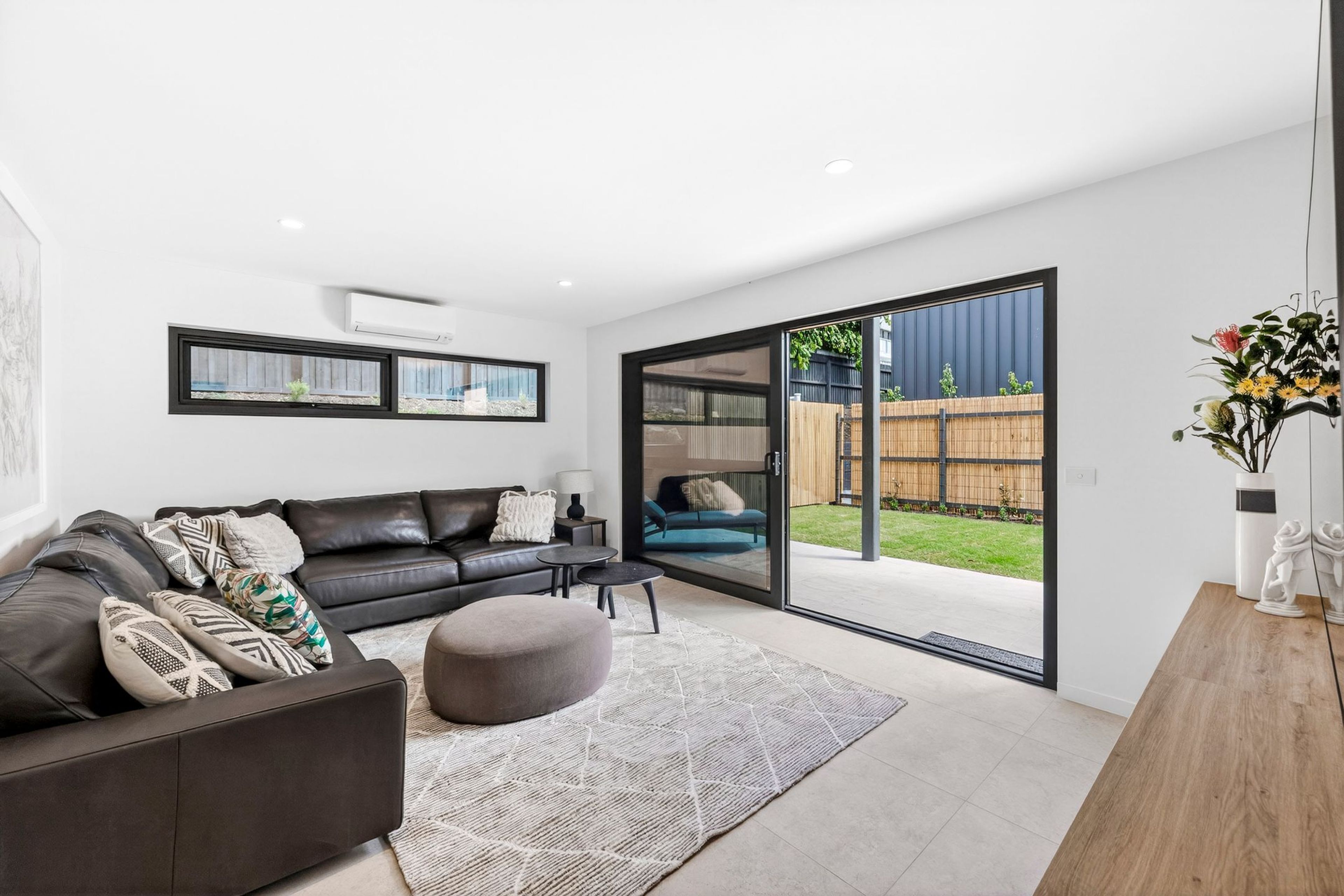 Un salon moderne avec un canapé en cuir noir, un pouf gris et un tapis à motifs. Une porte-fenêtre coulissante donne accès à un jardin avec une clôture en bois et de la verdure.