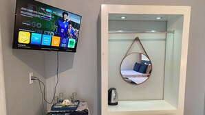 Habitación de hotel con televisión mostrando un canal deportivo, minibar, espejo y cama con lámpara colgante.