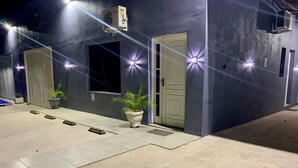 Exterior de un edificio de noche con fachada azul, una puerta blanca y plantas en macetas.