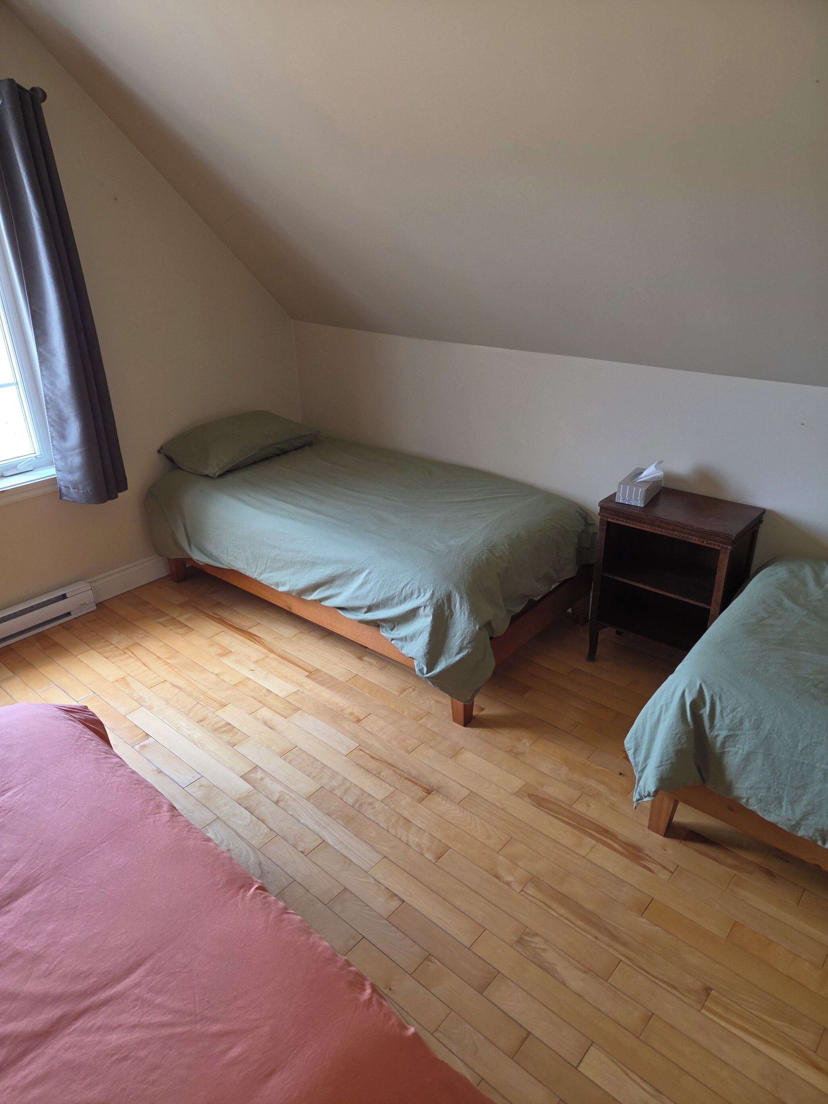 A small, single-room with a sloped ceiling, a bed with a green bedspread, a bedside table with a tissue box, and a large window with curtains.