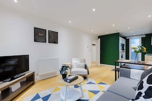 A modern living room with a flat-screen TV, a white armchair, a round coffee table, and a sofa. - Colourful 1BDR Canalside Flat in East London (Greater London)