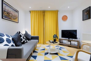 A living room with a sofa, a coffee table, a TV, and yellow curtains. - Colourful 1BDR Canalside Flat in East London (Greater London)