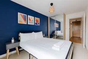 A neatly made bed with white linens, a bedside table with a lamp, and a wall with abstract art. - Colourful 1BDR Canalside Flat in East London (Greater London)