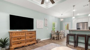A spacious living room with a large flat-screen TV, wooden chest of drawers, a dining area with a table and chairs, and a kitchen with white cabinets and a stainless steel refrigerator. - Cozy 2 Bedroom Atlas Cottage near Milton FL (Milton)