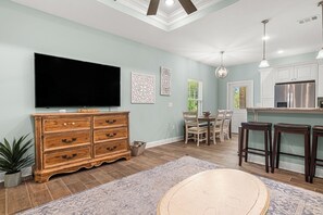 Ein geräumiges Wohnzimmer mit einem großen Flachbildfernseher, einer Holzkistenkommode, einem Essbereich mit Tisch und Stühlen sowie einer Küche mit weißen Schränken und einem Edelstahl-Kühlschrank.