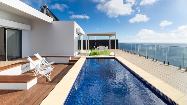 A modern rooftop pool with a clear view of the ocean, a covered seating area, and a wooden deck.