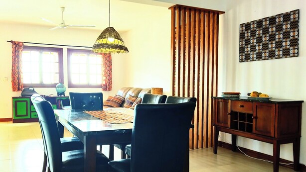 A dining area with a table, chairs, a sofa, a ceiling fan, a hanging light fixture, a patterned wall hanging, and a wooden cabinet.